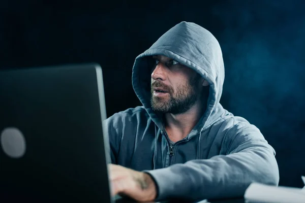 El hacker en una chaqueta gris con una capucha está sentado en el portátil y mirando a un lado — Foto de Stock