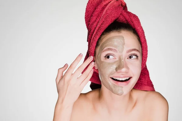 En lycklig ung flicka med vita tänder, med en röd handduk på huvudet, tillämpas en användbar lermask på halva ansiktet — Stockfoto
