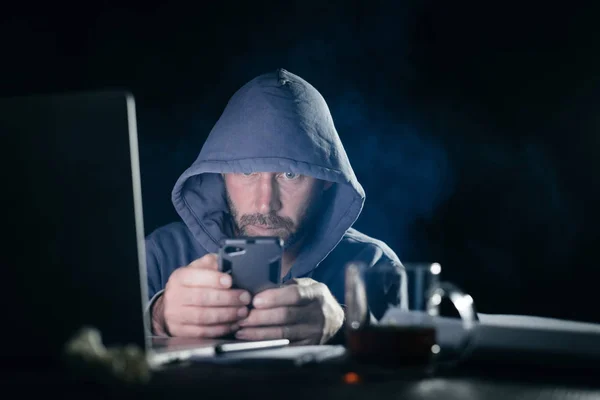 Adulto barbudo homem esconde seu rosto sob o capô, no escuro, um hacker, detém um smartphone — Fotografia de Stock