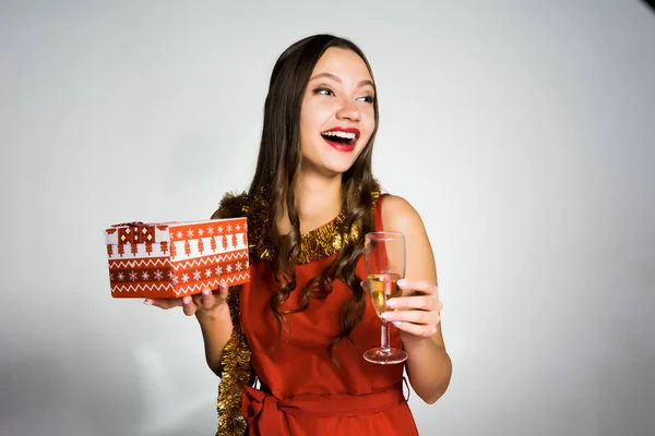 Счастливая молодая девушка в красном платье, с золотой мишурой на шее празднует Новый год, держа в руках подарок и бокал шампанского — стоковое фото