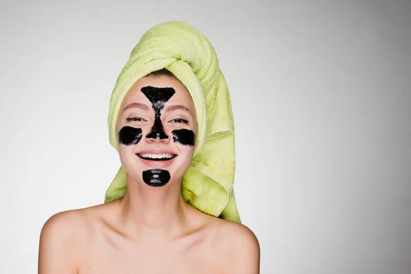 Laughing girl with a green towel on her head, on her face a black cleansing mask — Stock Photo, Image