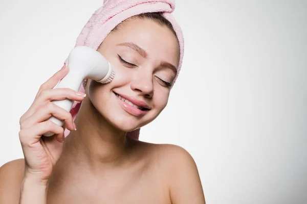 Happy smiling girl with a pink towel on her head doing a deep cleansing of her face with an electric brush — Stock Photo, Image