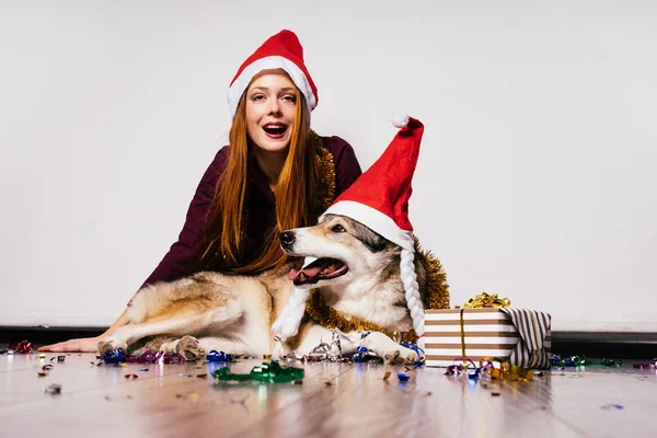 Счастливая рыжеволосая девушка, сидящая на полу с собакой в красных шапках в ожидании нового 2018 года — стоковое фото