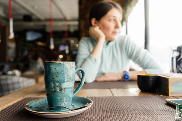 Macro de una hermosa taza y plato, en un café, una chica mira pensativamente por la ventana —  Fotos de Stock