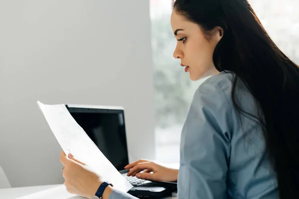 Giovane donna d'affari dai capelli neri che lavora su un computer portatile in possesso di documenti in mano — Foto Stock