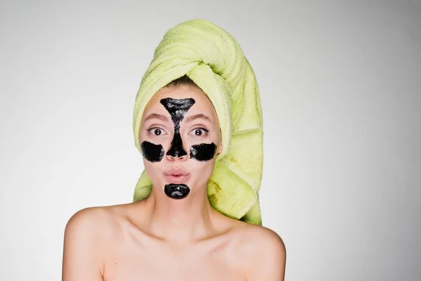 Ένα αστείο κορίτσι με μια πράσινη πετσέτα στο κεφάλι της εφαρμόζεται ένα μαύρο καθαρισμού μάσκα στις προβληματικές περιοχές στο πρόσωπό της — Φωτογραφία Αρχείου