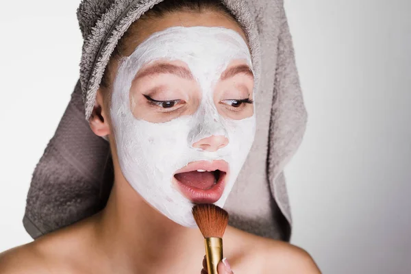 Ελκυστική κοπέλα με μια πετσέτα στο κεφάλι της, βάζει μια λευκή μάσκα στο πρόσωπό της με ένα πινέλο — Φωτογραφία Αρχείου
