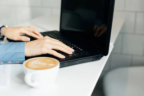 Девушка фрилансер в синей рубашке работает на ноутбуке, рядом кружка с ароматным кофе — стоковое фото