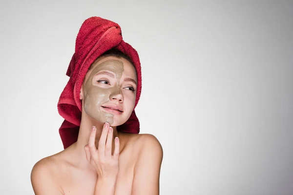 Gyönyörű lány, egy piros törülközőt a fején élvezi spa kezelések, agyag hasznos maszk a fél arca — Stock Fotó