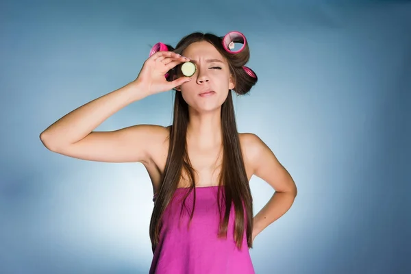 一个滑稽的女孩拿着一块黄瓜在眼睛附近, 滋润皮肤, 在卷发的头 — 图库照片