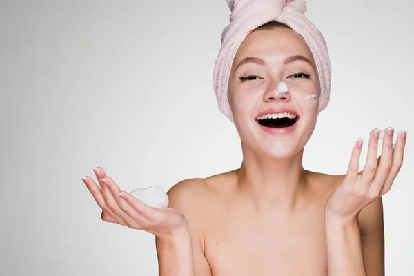 Смеется привлекательная девушка с полотенцем на голове, нанося увлажняющий крем на лицо — стоковое фото