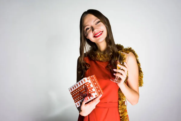 Happy smiling girl in a red dress and with a gold tinsel around her neck celebrating the new year 2018 and christmas, holding in her hand a gift and champagne — Stock Photo, Image