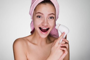 derin yüz cilt temizliği için elektrikli bir fırça tutan başında pembe havlu ile neşeli gülen kız