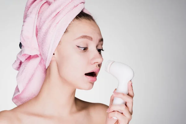 Dívka s růžový ručník na hlavě drží Elektrický kartáč v dlaních pro hluboké čištění obličeje, vypadá překvapeně — Stock fotografie