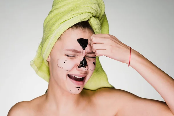 Ένα νεαρό κορίτσι με μια πράσινη πετσέτα στο κεφάλι της μια μαύρη μάσκα καθαρισμού αφαιρεί από το πρόσωπό της, να πονάει — Φωτογραφία Αρχείου