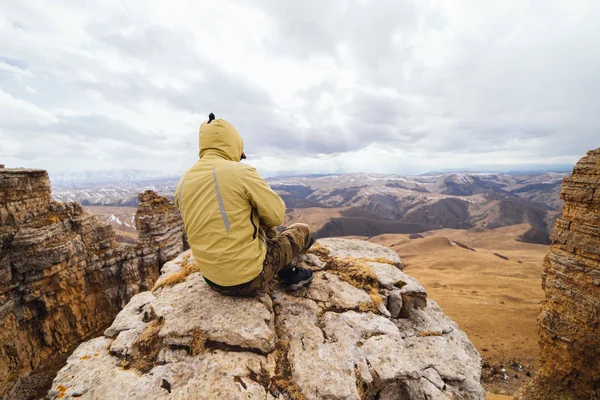 A male traveler in a yellow jacket sits on the edge of the mountain and enjoys nature and clean air — Stock Photo, Image