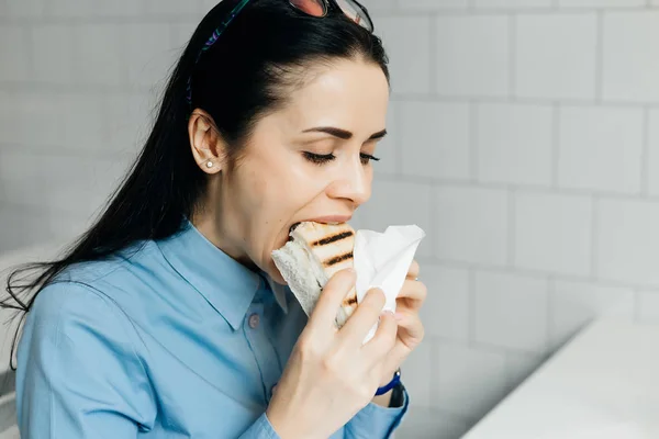 Молода брюнетка дівчина в блакитній сорочці охоче їсть бутерброд в кафе — стокове фото