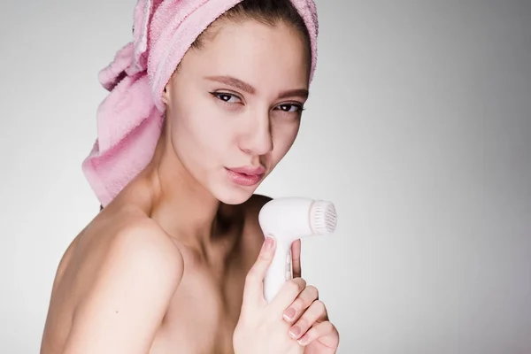 Jovem menina bonita com uma toalha rosa na cabeça detém uma escova elétrica especial para limpeza profunda de seu rosto — Fotografia de Stock