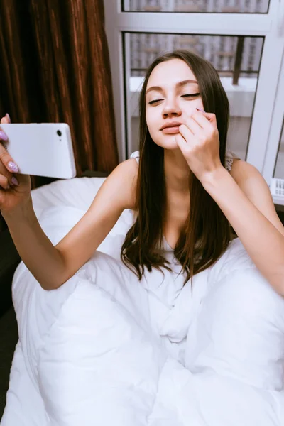 Schläfriges süßes Mädchen ist gerade aufgewacht, sitzt im Bett und macht Selfie — Stockfoto