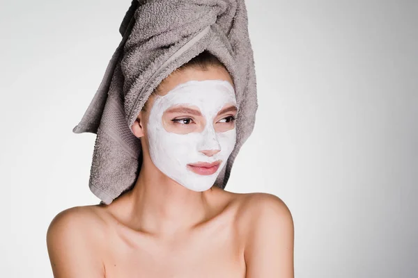 Ένα νεαρό κορίτσι με μια πετσέτα στο κεφάλι της εφαρμόζεται μια λευκή μάσκα για να καθαρίσει το πρόσωπό της — Φωτογραφία Αρχείου