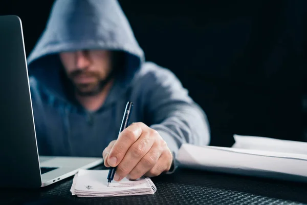 a young guy hacker in a sweatshirt with a hood sits behind a laptop and holds in his hand a pen with a notebook