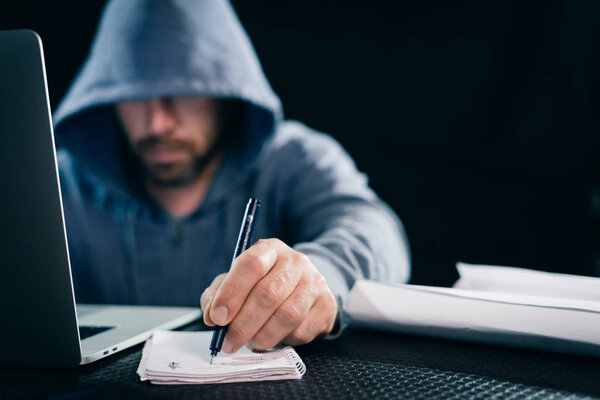 a young guy hacker in a sweatshirt with a hood sits behind a laptop and holds in his hand a pen with a notebook