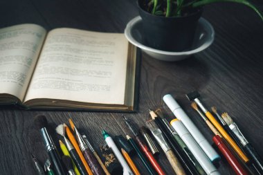 ahşap yüzeyde birçok kurşun kalem, açık bir kitap ve bir saksı bitki vardır