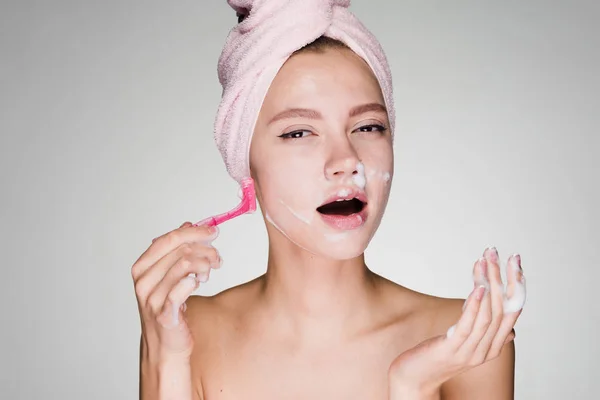 Divertida joven con una toalla rosa en la cabeza afeitándose la cara con una navaja rosa como un hombre — Foto de Stock