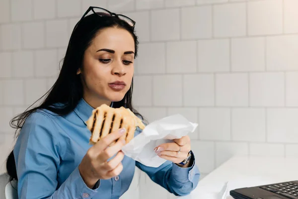 Мила брюнетка студентка в синій сорочці їсть бутерброд в кафе перед заняттям — стокове фото