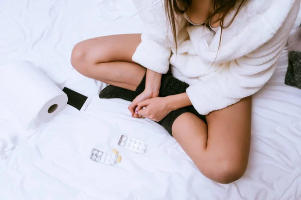 Uma menina em um vestido de vestir branco terry e meias quentes ficou doente, senta-se na cama, ao lado de um monte de comprimidos — Fotografia de Stock