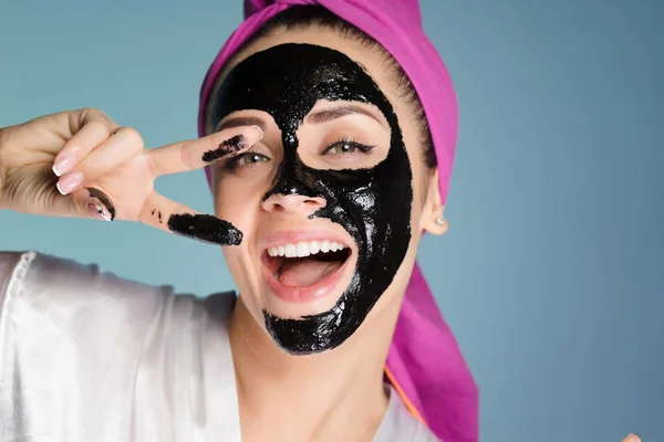 Uma menina sorridente feliz com dentes brancos, com uma toalha rosa na cabeça mostrando dois dedos, uma máscara de limpeza preta em seu rosto — Fotografia de Stock