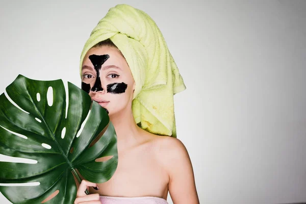 Una joven se cuida la cara, se aplica una máscara de limpieza negra en la cara, una toalla verde en la cabeza, un día de spa — Foto de Stock