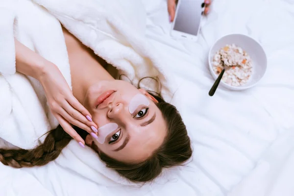 Giovane ragazza carina in una vestaglia di spugna bianca è sdraiato sul letto, accanto a un piatto di farina d'avena per la prima colazione — Foto Stock