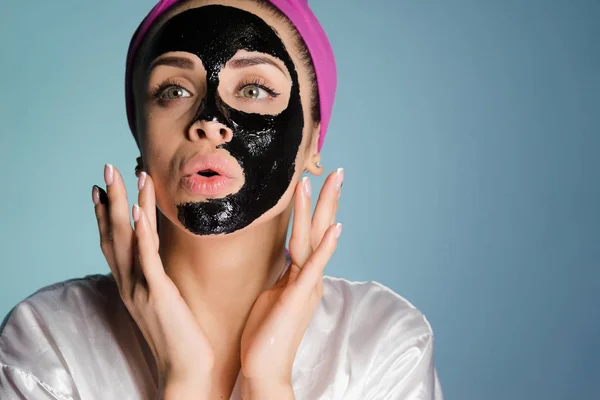 En rolig ung flicka med rosa handduk på hennes huvud vill se bra ut, sätter en svart cleansing mask på ansiktet — Stockfoto