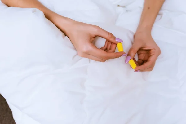 Dziewczyna siedzi na łóżku pod biały koc, w rękach żółte zatyczki do uszu przeciw hałas z ulicy — Zdjęcie stockowe