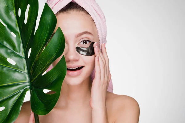 Smiling beautiful girl with a pink towel on her head holding a green leaf in her hand, under the eyes of black patches, spa procedures — Stock Photo, Image