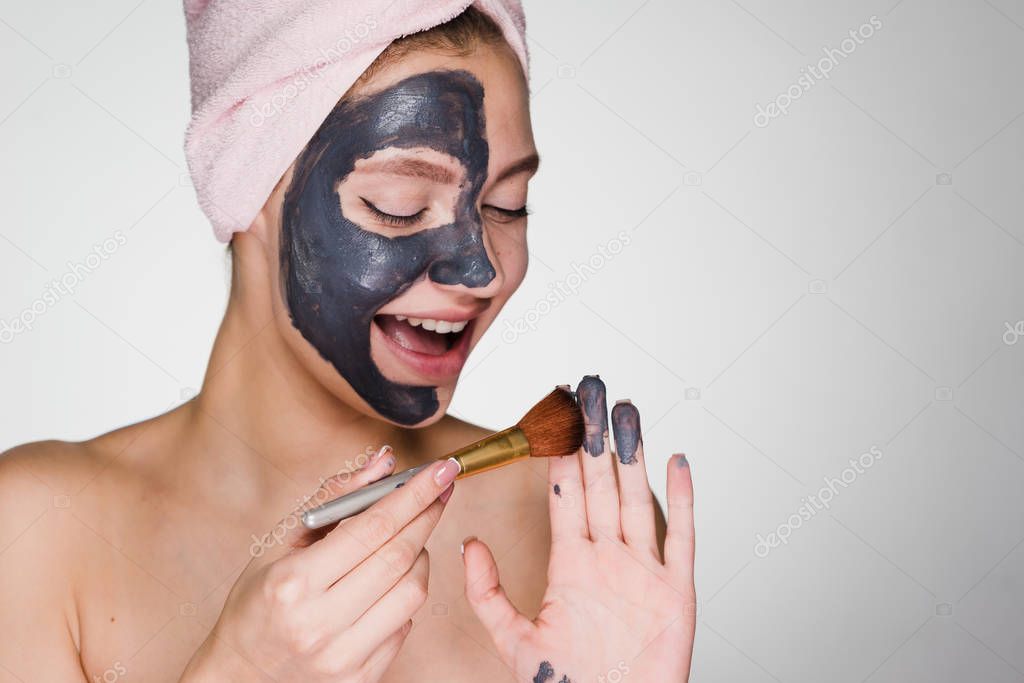 a smiling sweet girl with a pink towel on her head applies a useful clay mask on her face with a brush