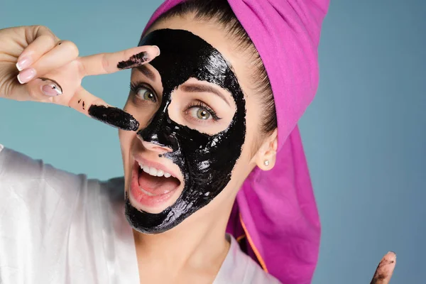 Ευτυχισμένος χαμογελαστό κορίτσι με μια ροζ πετσέτα στο κεφάλι της εφαρμόζεται μια μαύρη μάσκα κατά της ακμής στο πρόσωπό της, δείχνει δυο δάχτυλα — Φωτογραφία Αρχείου