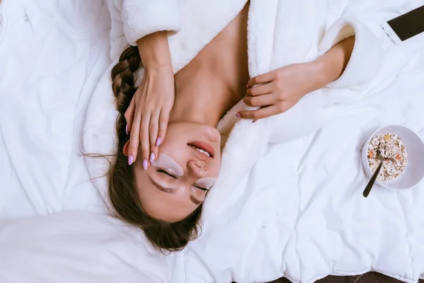 Jovem menina em um manto branco jaz na cama, no início da manhã, sob os patches olhos — Fotografia de Stock