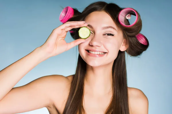 Usměvavá dívka s natáčky na hlavě drží kus okurky pro zvlhčení kůže na tváři — Stock fotografie