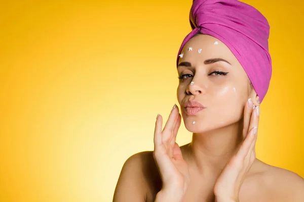 Giovane ragazza con un asciugamano rosa sulla testa applicare crema nutriente bianca sul suo viso, su uno sfondo giallo — Foto Stock