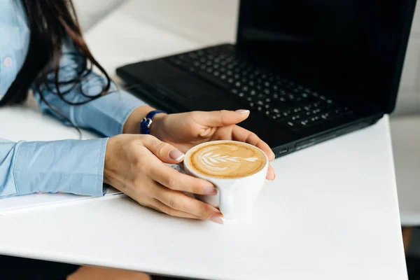 Una chica en una camisa azul bebe un café fragante en un café, cerca de un ordenador portátil para el trabajo — Foto de Stock