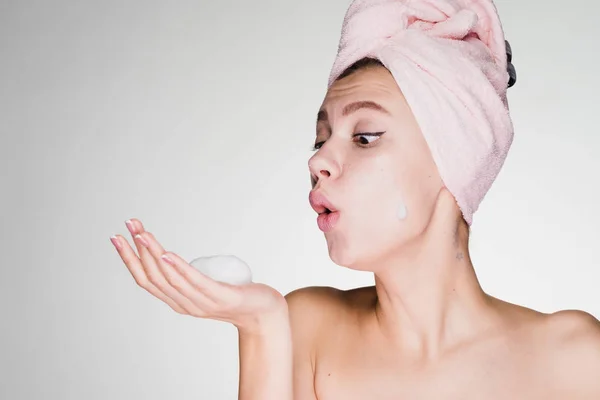 Funny funny girl with a pink towel on her head blowing on cleansing facial foam — Stock Photo, Image