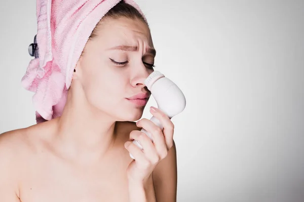 Sad young girl with a pink towel makes a deep cleansing of the skin on her face with an electric brush — Stock Photo, Image