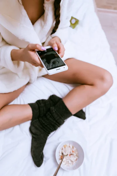 Dziewczyna w szlafrok biały frotte i ciepłe skarpety siedzi w łóżku i wykonywanie zdjęć z siebie — Zdjęcie stockowe