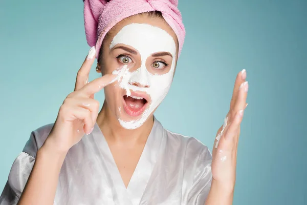 A funny surprised young girl with a pink towel puts on her face a white nutritious mask — Stock Photo, Image