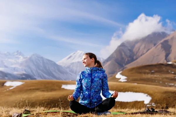 Lächelndes junges Mädchen in blauer Jacke meditiert vor dem Hintergrund der kaukasischen Berge in der Sonne — Stockfoto