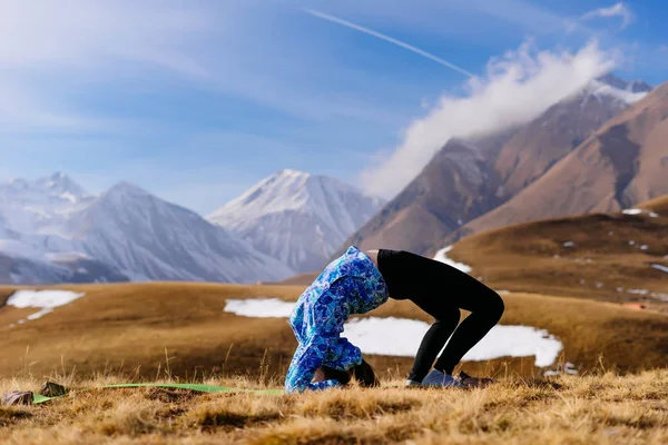 Спортивная девушка в синей куртке занимается йогой на фоне Кавказских гор — стоковое фото