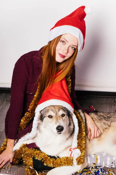 Рыжеволосая красивая девушка празднует Новый год со своей собакой, на голове красная шапка, как Санта-Клаус — стоковое фото