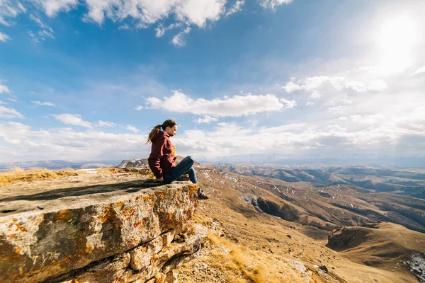 Active young girl sits on the edge of the cliff, enjoys the mountain air and the sun — Stock Photo, Image